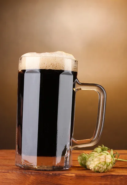 Birra scura in una tazza e luppolo verde su tavolo di legno su sfondo marrone — Foto Stock