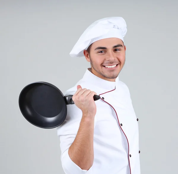 Chef profesional en uniforme blanco y sombrero, sosteniendo la sartén en sus manos, sobre fondo gris — Foto de Stock