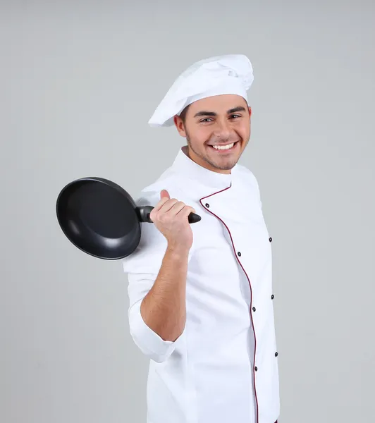 Профессиональный повар в белой форме и шляпе, держащий сковородку в руках, на сером фоне — стоковое фото