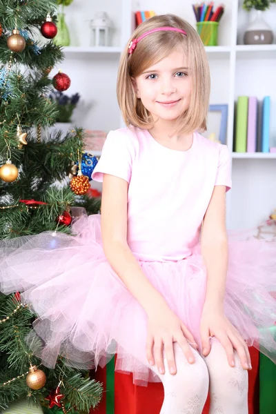 Little girl sitting on big present box near Christmas tree in room — Stock Photo, Image