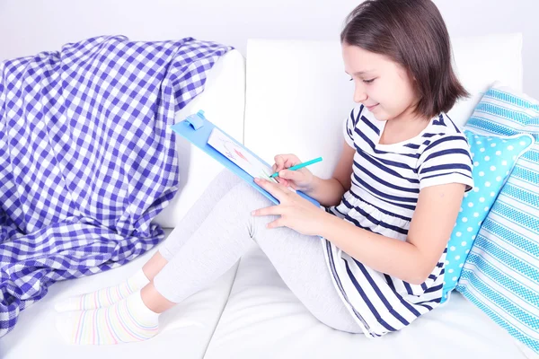 Beautiful little girl sitting on sofa and drawing picture, on home interior background