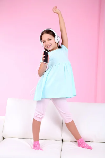 Beautiful little girl listening to music and dancing on sofa  and listening to music, on home interior background — Stock Photo, Image