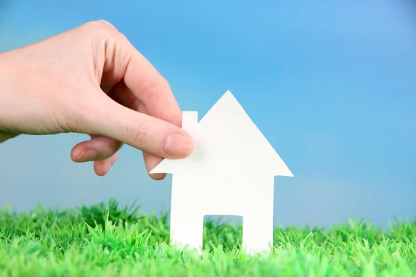 Little paper house in hand close-up, on green grass, on blue sky background — Stock Photo, Image