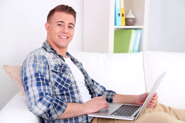 Bonito jovem sentado no sofá com laptop no quarto — Fotografia de Stock