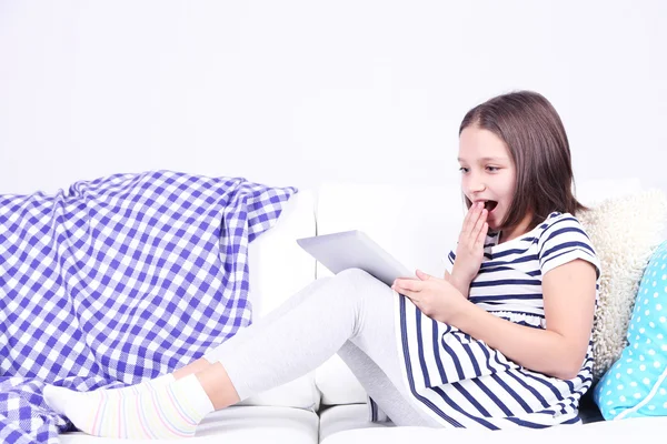 Hermosa niña sentada en el sofá con la tableta, en el fondo interior del hogar —  Fotos de Stock