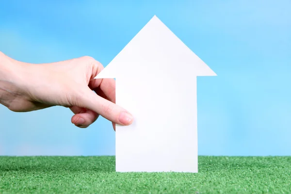 Little paper house in hand close-up, on green grass, on blue sky background — Stock Photo, Image