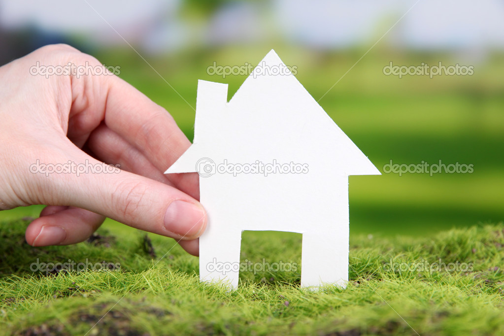 Little paper house in hand close-up, on green grass, on bright background