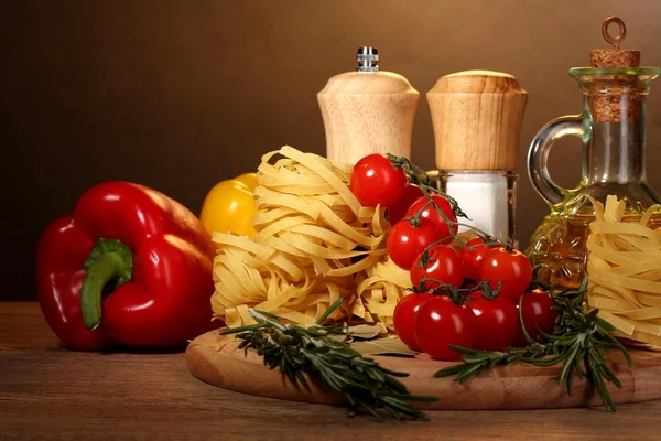Nudlar i skål, burk med olja, kryddor och grönsaker på träbord på brun bakgrund — Stockfoto