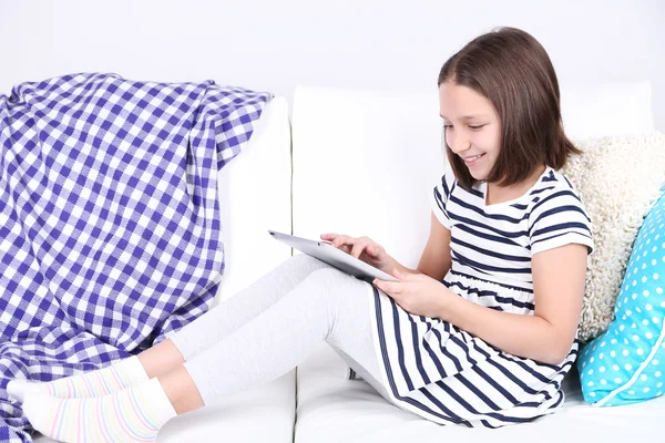 Menina bonita sentada no sofá com tablet, em casa fundo interior — Fotografia de Stock