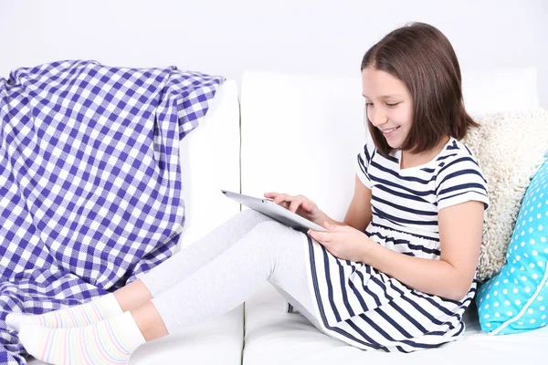 Beautiful little girl sitting on sofa with tablet, on home interior background — Stock Photo, Image