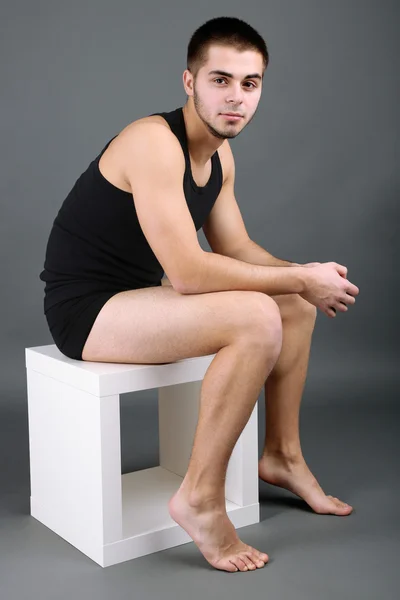 Pohledný mladý sedící na krychli na šedém pozadí — Stock fotografie