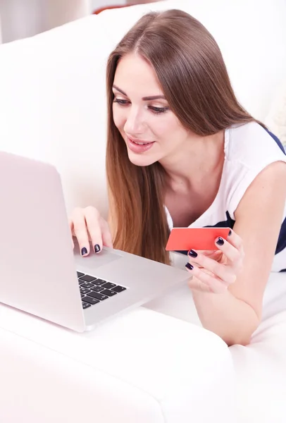 Jovem sentada com laptop no sofá e segurando cartão de crédito na mão, em casa — Fotografia de Stock