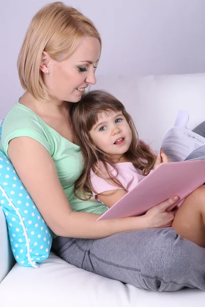 Pretty little girl reading book with mother on sofa on gray background — Stock Photo, Image
