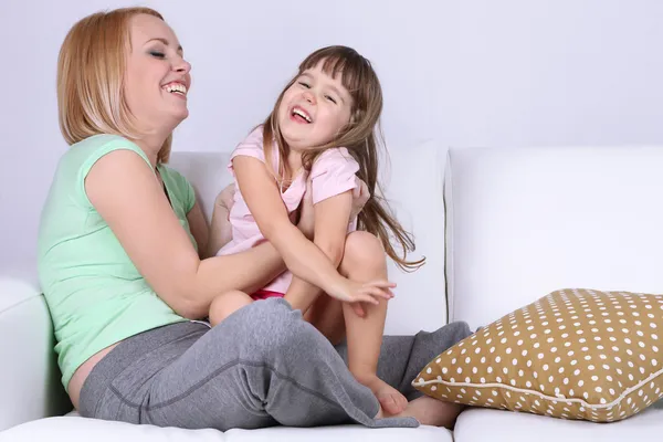 Hübsches kleines Mädchen sitzt mit Mutter auf Sofa auf grauem Hintergrund — Stockfoto
