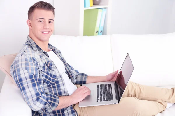 Schöner junger Mann sitzt auf Couch mit Laptop im Zimmer — Stockfoto