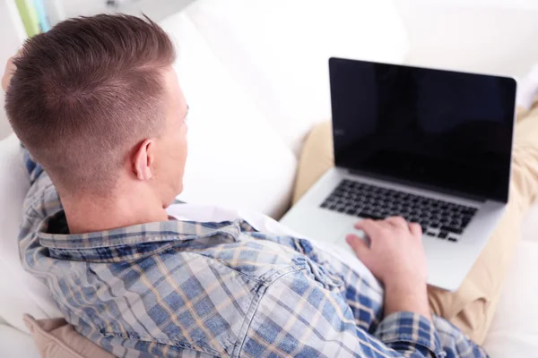 Przystojny młody mężczyzna siedzi na kanapie z laptopem w pokoju — Zdjęcie stockowe