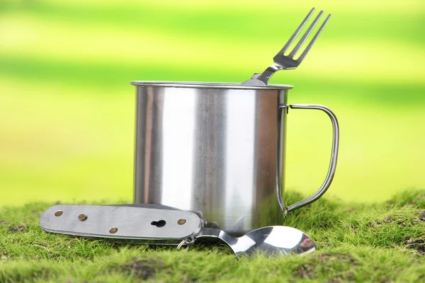 Metall mugg, sked och gaffel på grönt gräs, på natur bakgrund — Stockfoto