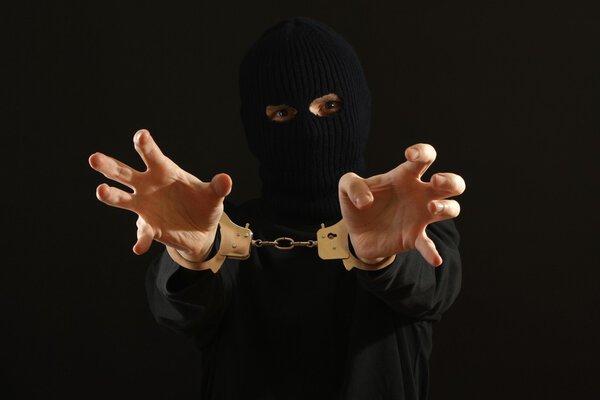 Angry bandit in black mask handcuffed isolated on black