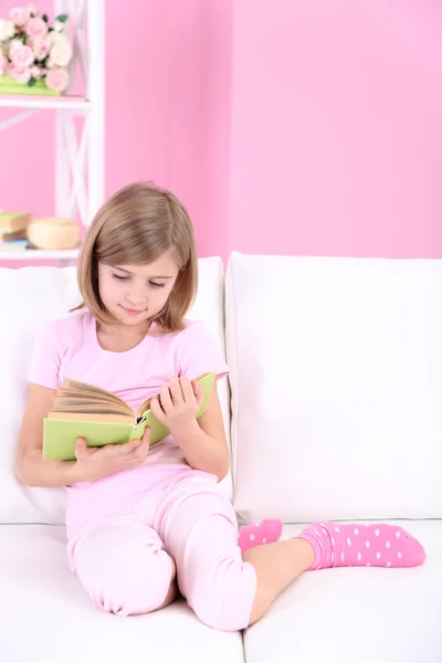 Niña leyendo libro sentado en el sofá en la habitación —  Fotos de Stock