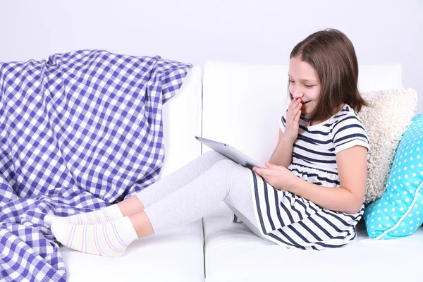 Beautiful little girl sitting on sofa with tablet, on home interior background — Stock Photo, Image