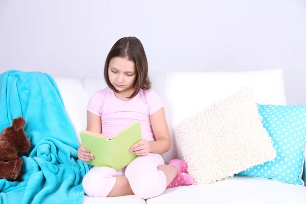 Bella bambina seduta sul divano con libro, su sfondo interno di casa — Foto Stock