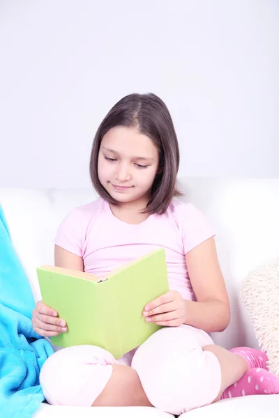 Hermosa niña sentada en el sofá con libro, en el fondo interior de casa —  Fotos de Stock