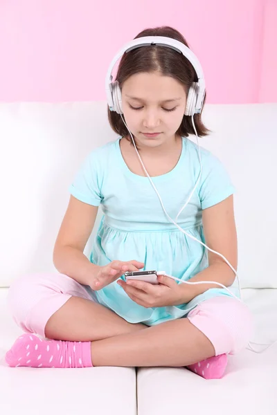 Beautiful little girl sitting on sofa  and listening to music, on home interior background — Stock Photo, Image