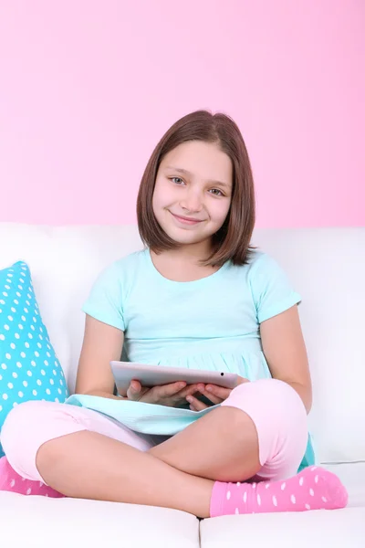 Hermosa niña sentada en el sofá con la tableta, en el fondo interior del hogar —  Fotos de Stock