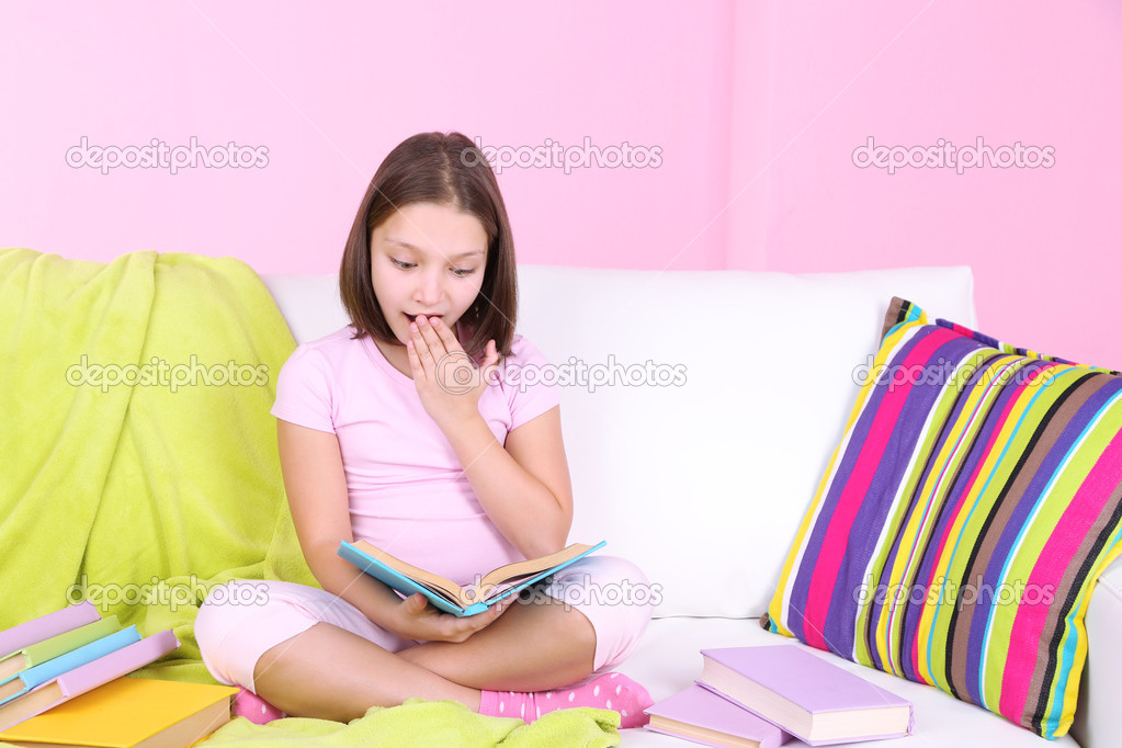 Beautiful little girl sitting on sofa with  book, on home interior background