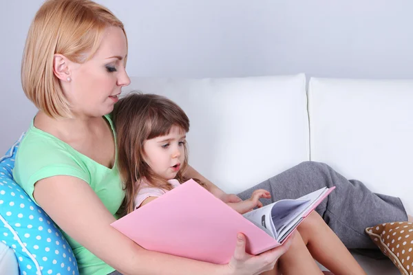 Linda niña leyendo libro con madre en sofá sobre fondo gris —  Fotos de Stock