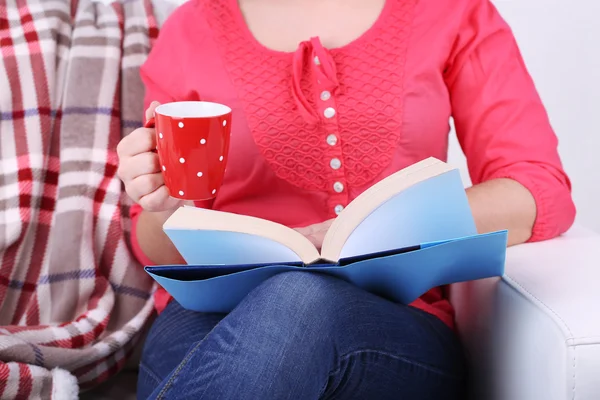Mulher sentada no sofá, lendo livro e beber café ou chá, close-up — Fotografia de Stock