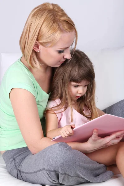 Pretty little girl reading book with mother on sofa on gray background — Stock Photo, Image