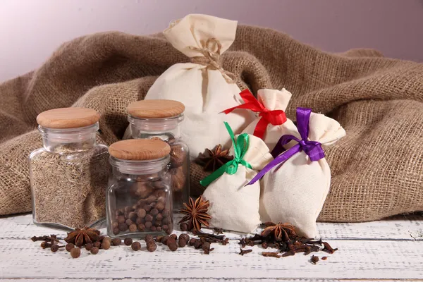 Sacks and glass jars full with spices, on wooden table, on sackcloth background — Stock Photo, Image