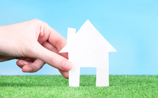 Little paper house in hand close-up, on green grass, on blue sky background — Stock Photo, Image