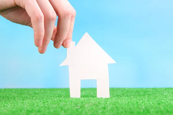 Little paper house in hand close-up, on green grass, on blue sky background — Stock Photo, Image