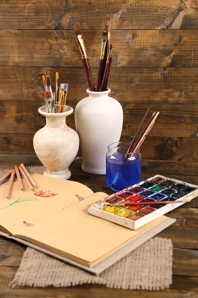 Composition with colorful watercolors, brushes in vase and sketcher on wooden background — Stock Photo, Image
