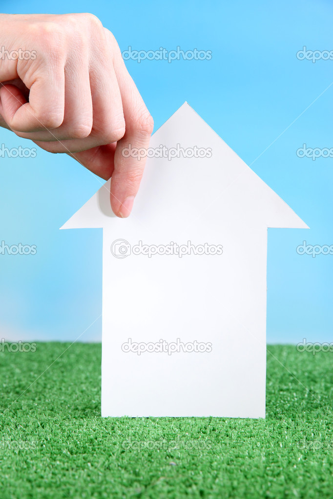 Little paper house in hand close-up, on green grass, on blue sky background
