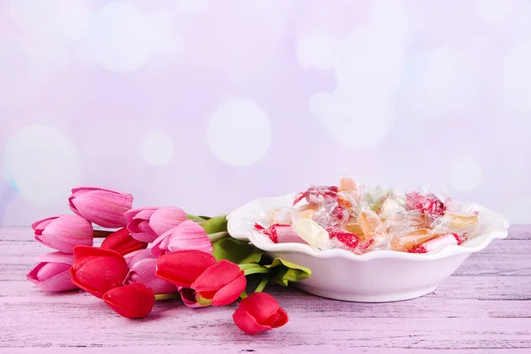 Sabrosos caramelos en tazón con flores en la mesa sobre fondo brillante —  Fotos de Stock