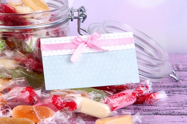 Sabrosos caramelos en frasco con tarjeta en la mesa sobre fondo brillante — Foto de Stock