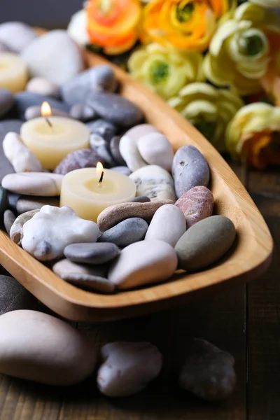 Tigela de madeira com pedras de spa e velas na mesa de madeira, no fundo flores — Fotografia de Stock