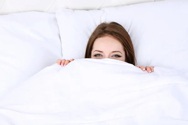 Jovem bela mulher olha para fora sob capas na cama close-up — Fotografia de Stock