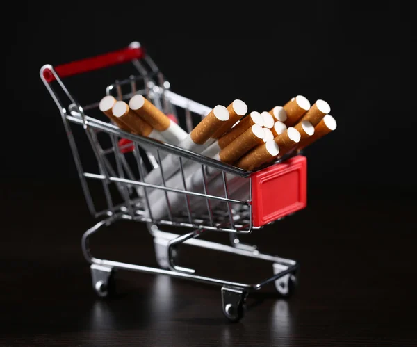 Cigarettes in shopping cart on wooden table on dark background — Stock Photo, Image
