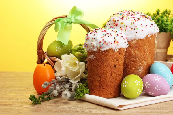 Belos bolos de Páscoa, ovos coloridos na cesta e velas na mesa de madeira no fundo amarelo — Fotografia de Stock