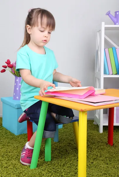 Little girl reads book sitting at table in room — Stock Photo, Image