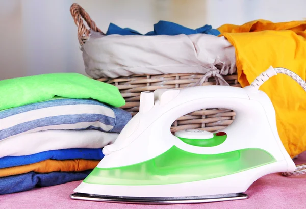 Iron and pile of colorful clothes and basket on table on bright background — Stock Photo, Image