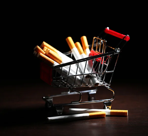 Cigarettes in shopping cart on wooden table on dark background — Stock Photo, Image