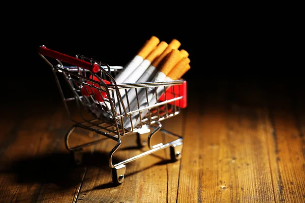 Cigarety v nákupním košíku na dřevěný stůl na tmavém pozadí — Stock fotografie
