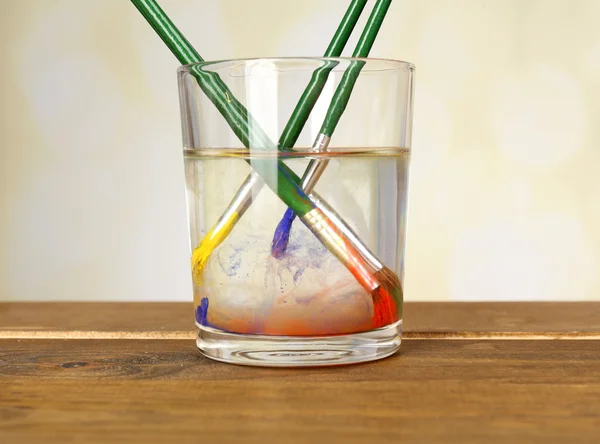 Brushes with color paint in glass of water, on wooden table — Stock Photo, Image