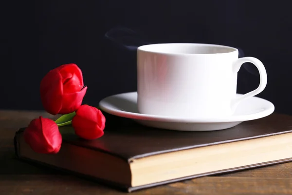 Tasse heißen Kaffee auf Buch mit Blumen auf Tisch auf dunklem Hintergrund — Stockfoto