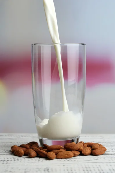 O leite de amêndoa é derramado em vidro, na tabela de madeira da cor, no fundo claro — Fotografia de Stock